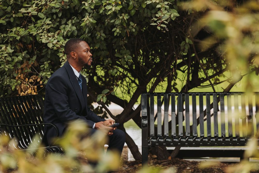 Man in suit sitting on park bench surrounded by green trees and foliage on a sunny day.