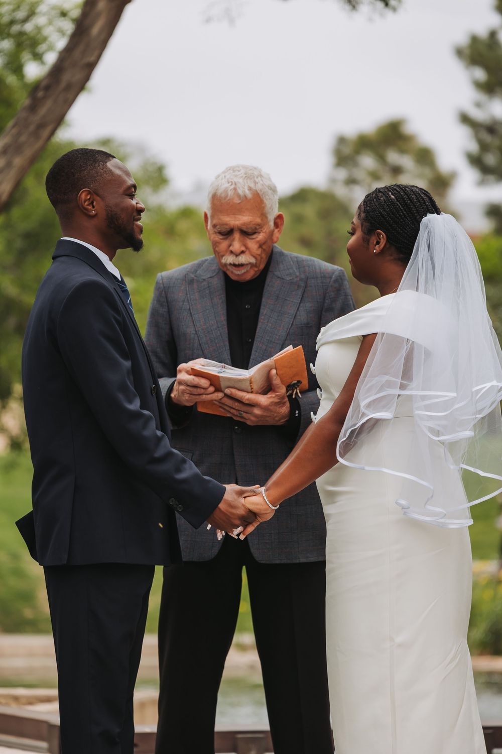 A bride and groom hold hands during an outdoor wedding ceremony while an officiant reads from a book.