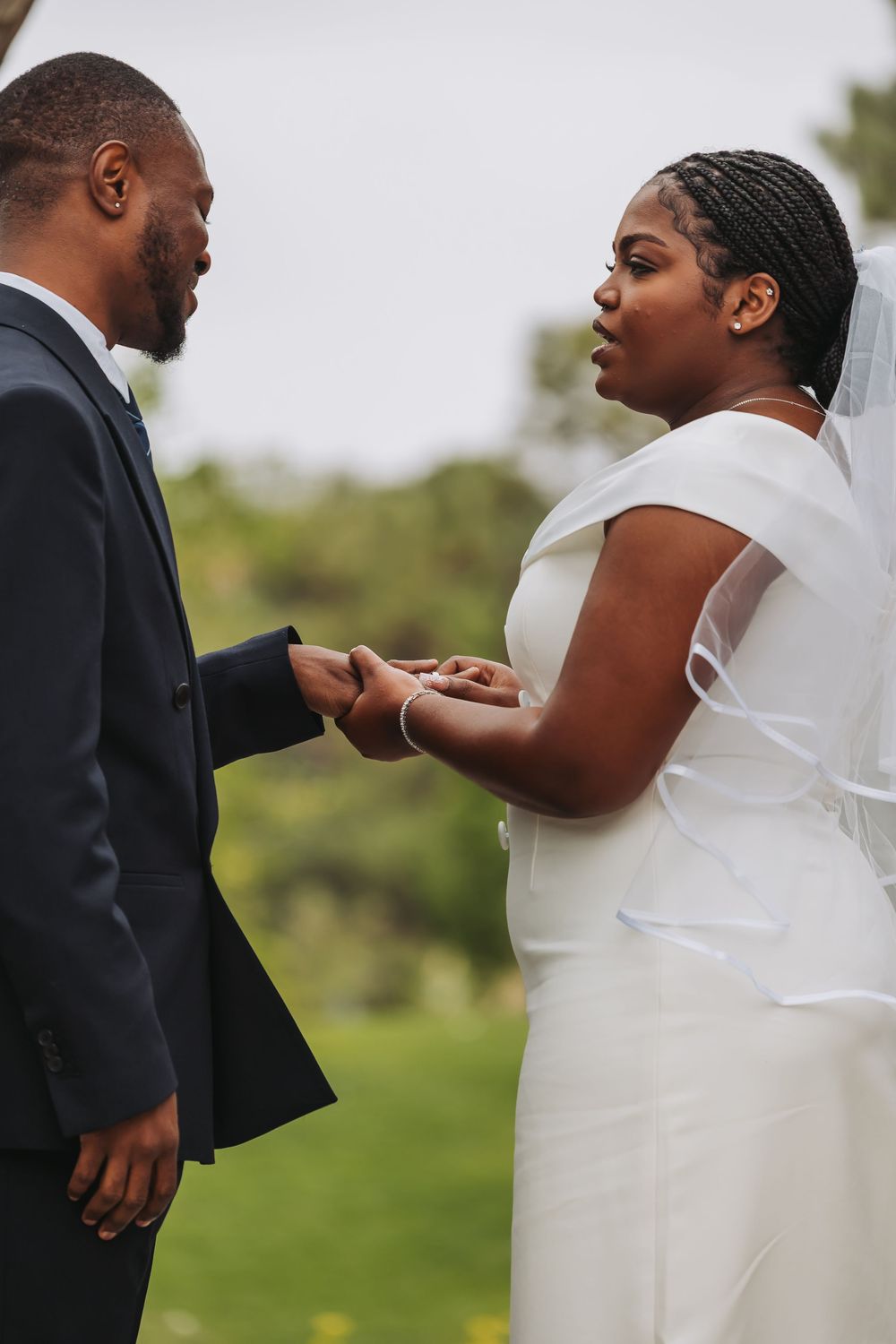 Bride and groom holding hands during outdoor wedding ceremony, exchanging vows in elegant white dress and dark suit.
