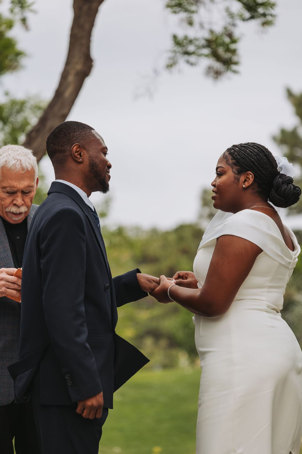 Bride placing wedding ring on groom's finger during outdoor ceremony, officiant standing nearby.