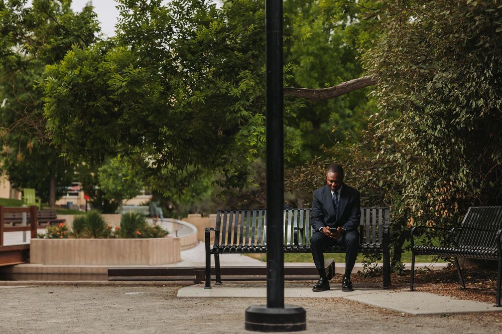 A businessman in a suit sits on a park bench looking at his phone, surrounded by lush green trees.