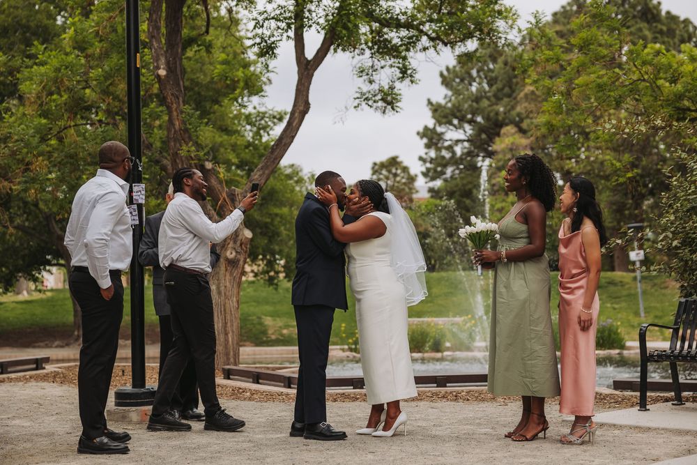 A joyful outdoor wedding ceremony with a couple kissing, surrounded by family in a lush park setting.