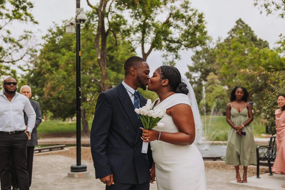 A bride and groom share a kiss outdoors at a park wedding ceremony, with guests watching in the background.