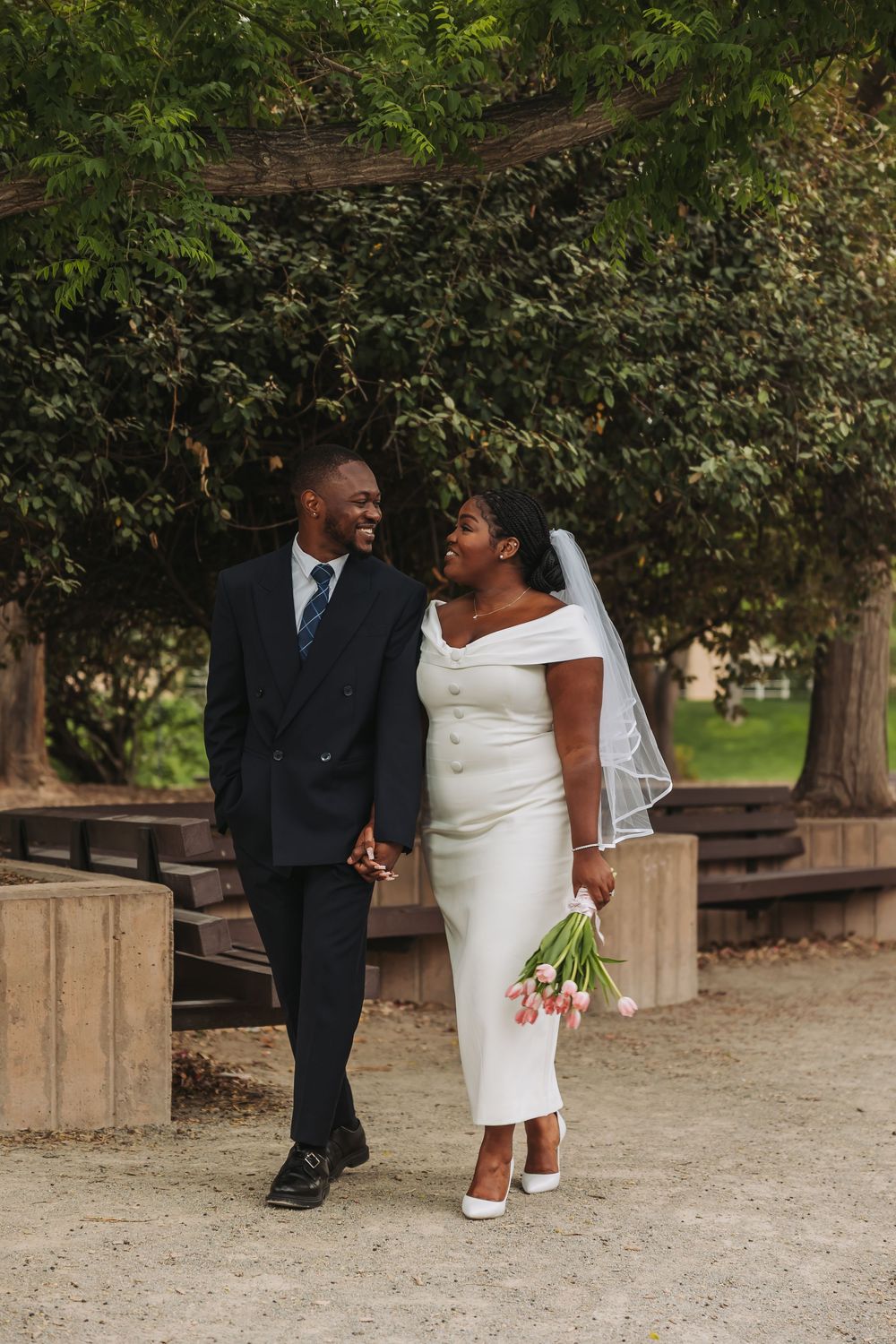 A joyful Black couple walks hand-in-hand in a park, bride in white midi dress with veil, groom in navy suit.