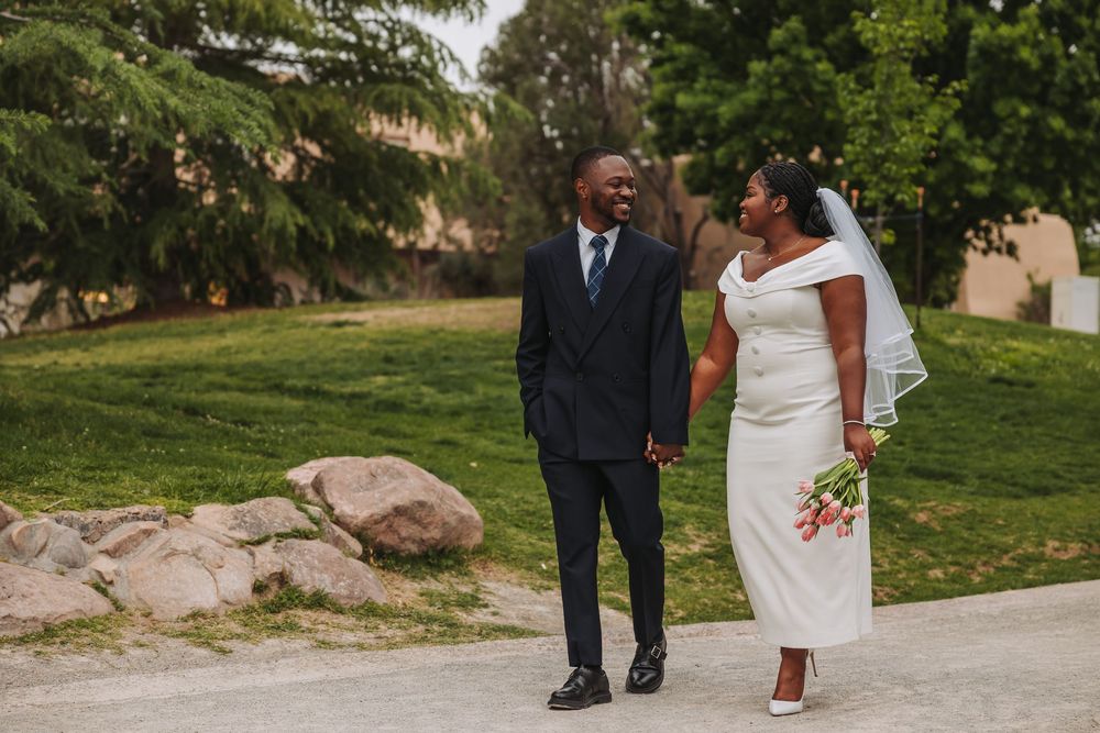 A newlywed Black couple holds hands and smiles at each other outdoors, bride in white dress with bouquet, groom in dark suit.