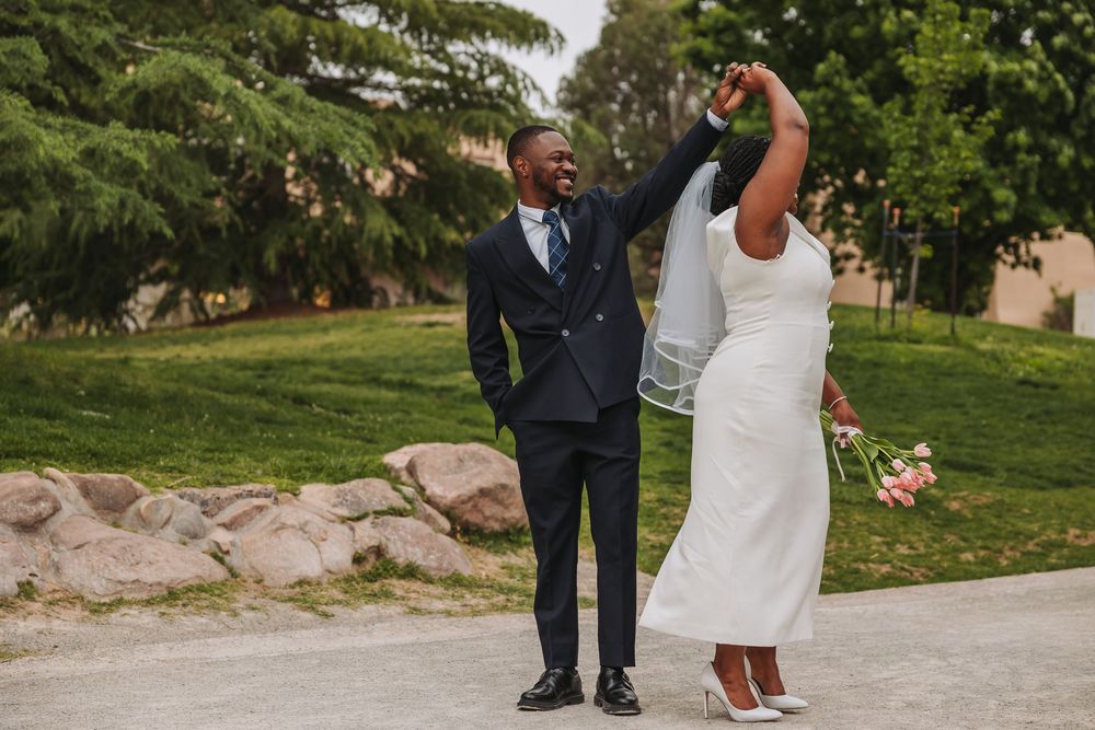 Joyful newlywed couple celebrating outdoors, groom in navy suit raising bride's hand as she twirls in white wedding dress.