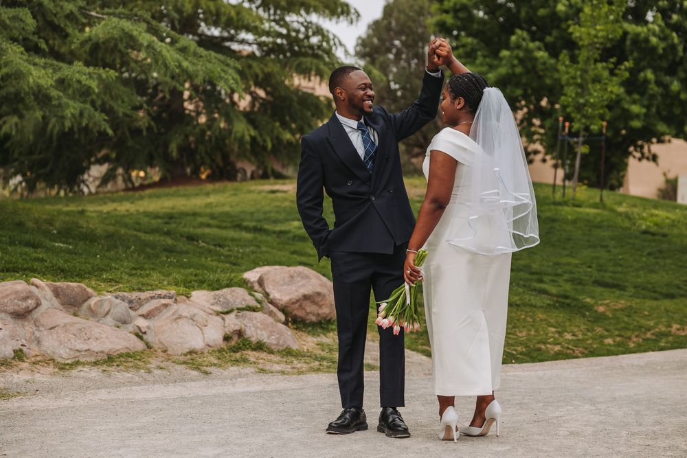 Happy Black groom raises bride's hand as they walk outdoors, bride holding white flowers in elegant wedding attire.