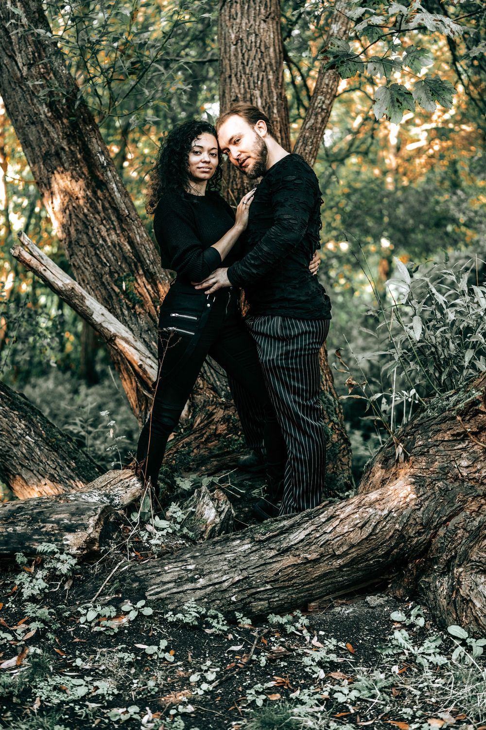 Interracial couple embracing romantically in a forest, both wearing black outfits, standing on fallen logs among trees.