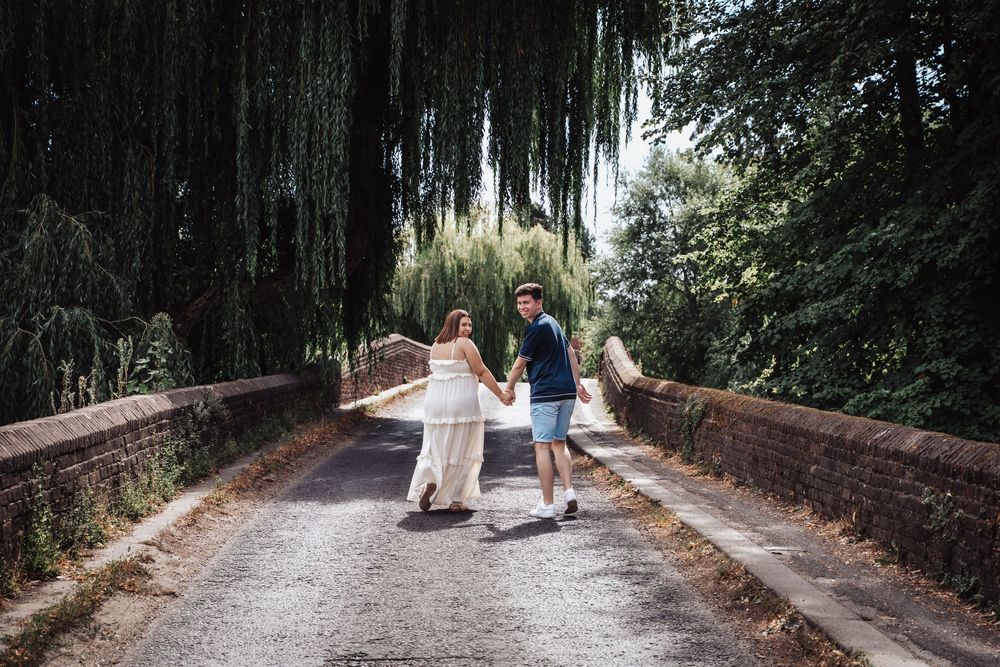 Couple holding hands walking on a bridge path surrounded by lush green weeping willow trees.