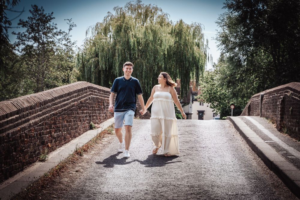Couple holding hands walking on a brick bridge surrounded by willow trees on a sunny day.