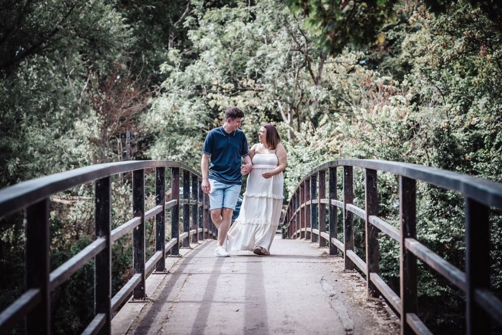 Couple walking on a wooden bridge surrounded by green trees in a romantic outdoor maternity photo.