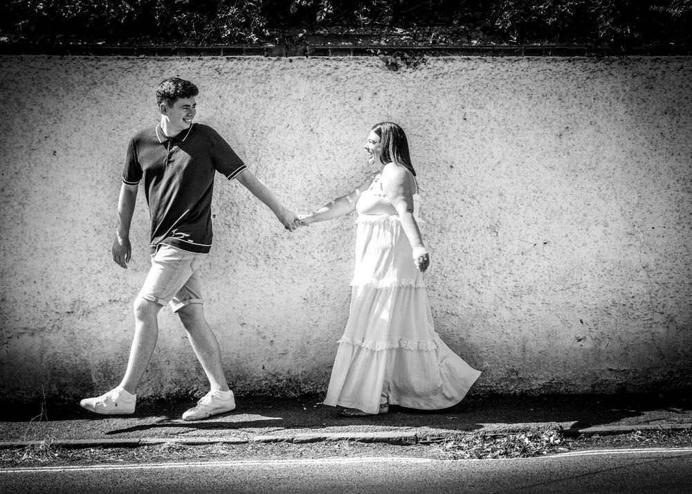 Black and white photo of a couple holding hands walking along a wall, woman in white dress, man in casual attire.