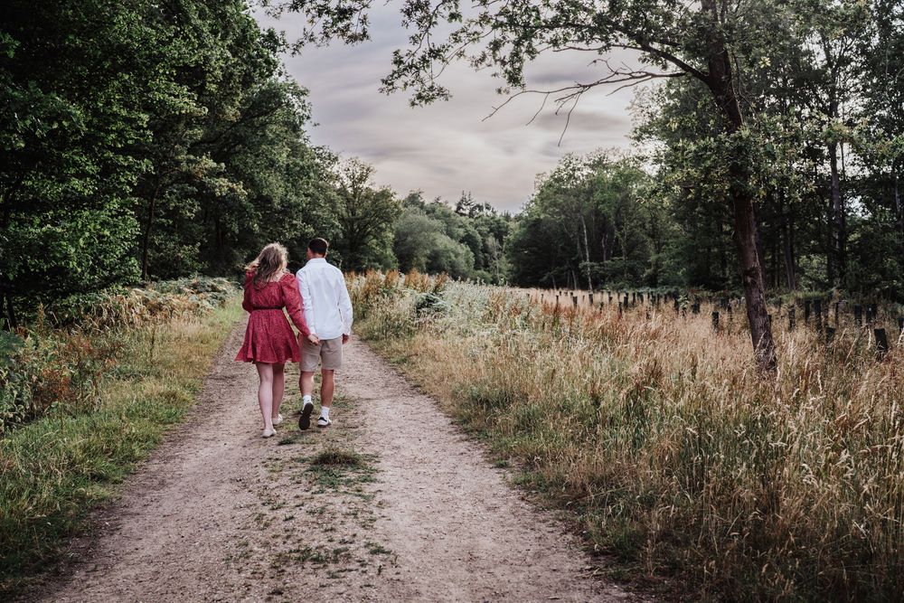 Couple walking hand in hand down a scenic dirt path surrounded by lush green trees and golden fields.