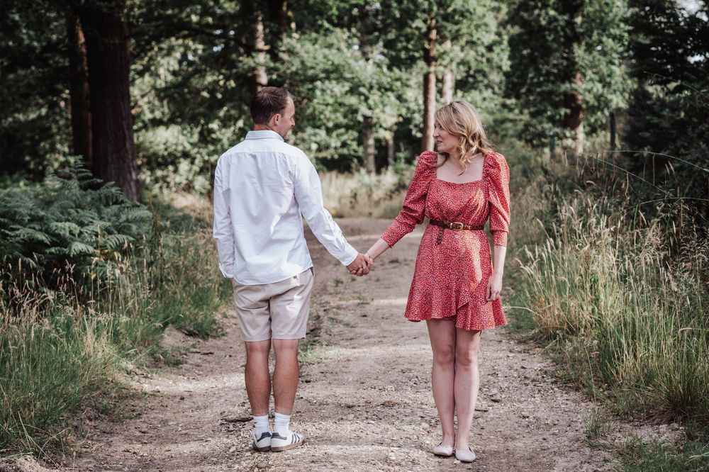 Couple holding hands walking on a dirt path through a lush green forest, woman in red dress, man in white shirt.