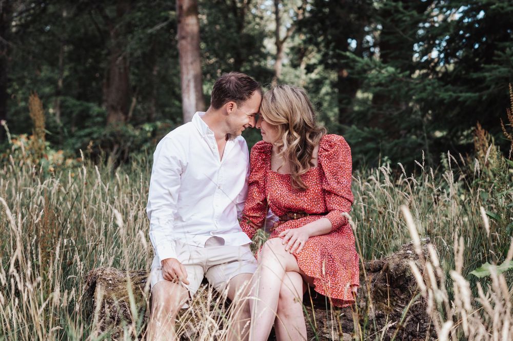 Couple sitting close together in a field of tall grass, sharing a romantic moment with trees in the background.