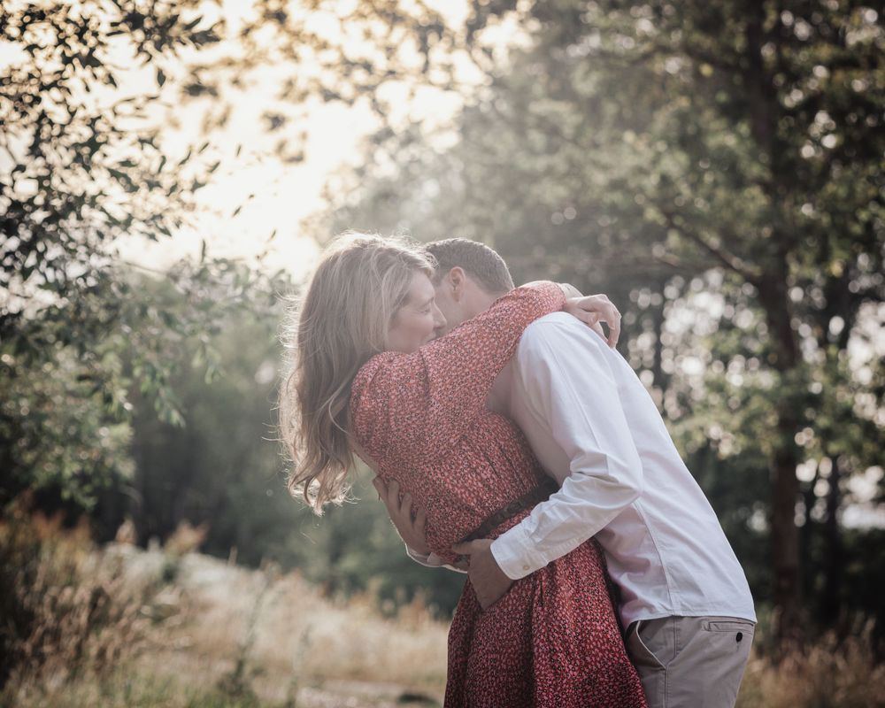 A couple embraces and kisses in a sunlit forest path, the woman wearing a red dress in a romantic outdoor setting.