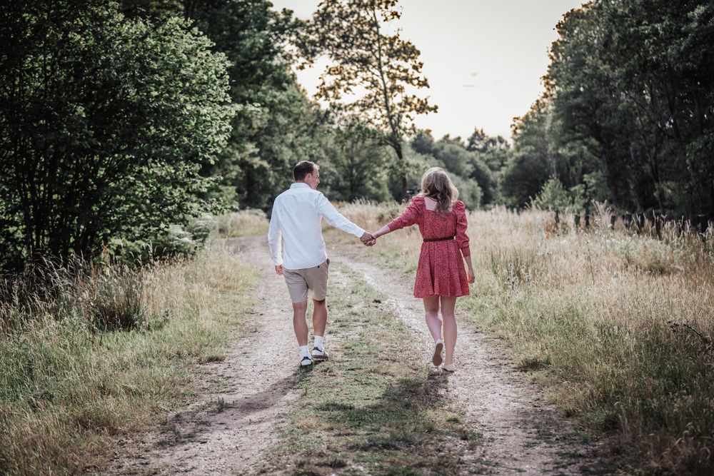 Couple holding hands walking down a scenic dirt path surrounded by lush green trees and tall grass at sunset.