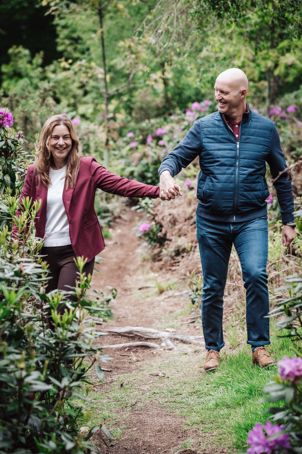 A smiling couple holds hands while walking along a garden path surrounded by lush greenery and purple flowers.
