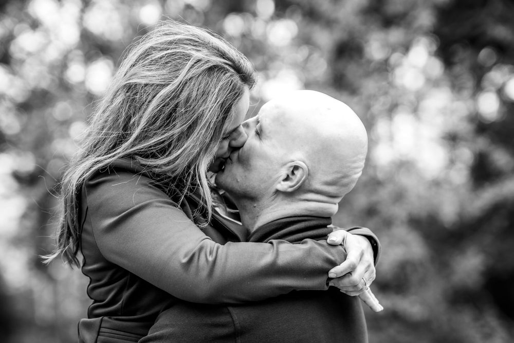 Black and white photo of a couple sharing a romantic kiss outdoors, surrounded by blurred trees.