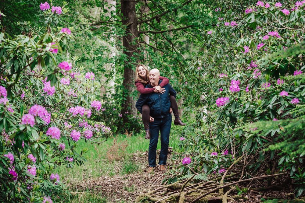 Couple sharing a piggyback ride among blooming pink rhododendrons in a lush green forest path.