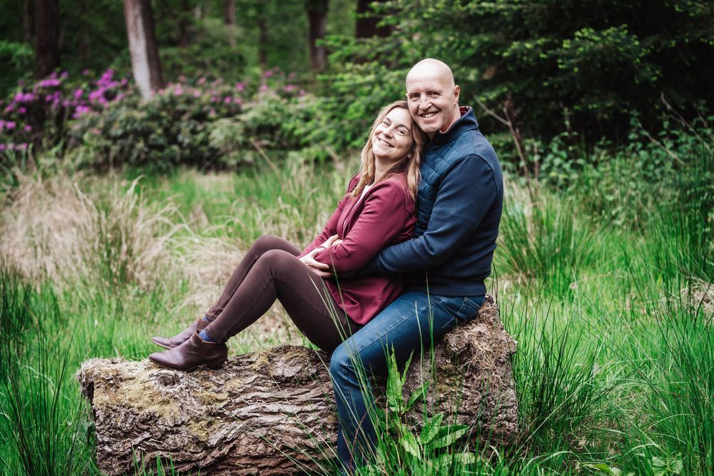 Couple sitting on a log in a lush green forest, smiling and embracing in a romantic outdoor portrait.