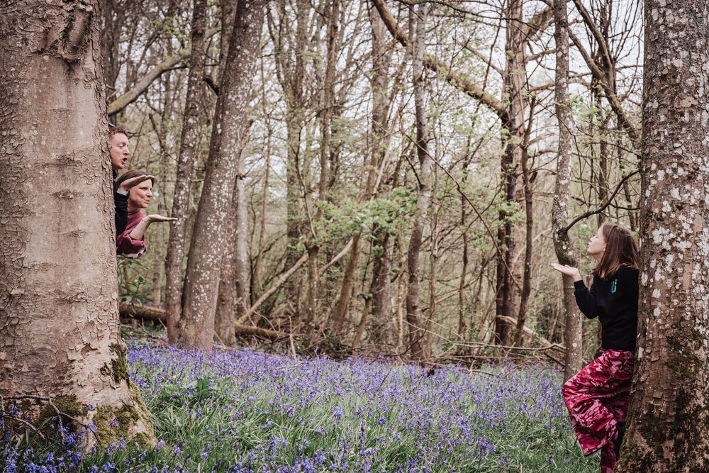 Two people playing among bluebell-covered forest floor, one peeking from behind a tree trunk in a woodland setting.