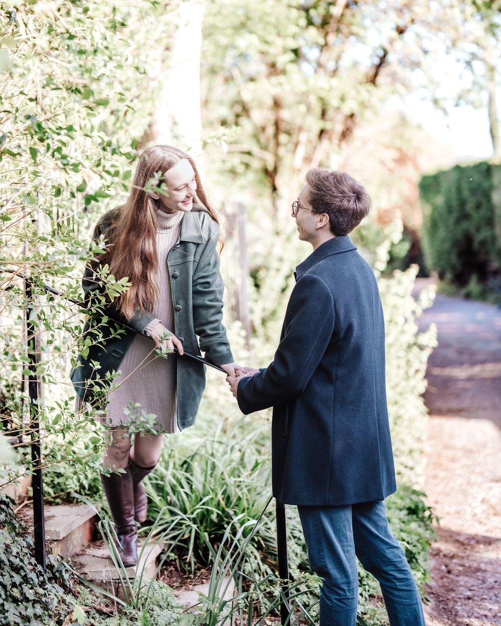 A couple holds hands in a lush garden, the woman in a green coat and boots, the man in a navy coat.