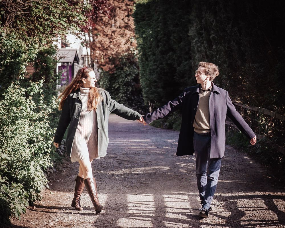 A couple holds hands while walking apart on a sunlit garden path surrounded by lush green hedges.