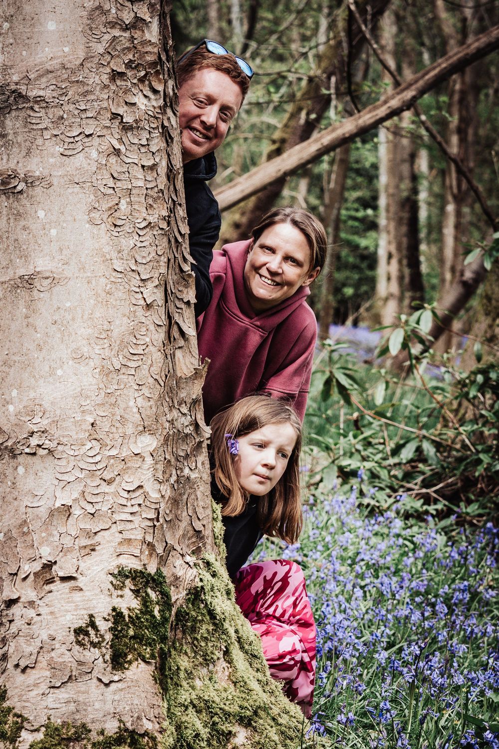 Family of three peeking from behind a mossy tree trunk in a bluebell woodland forest.