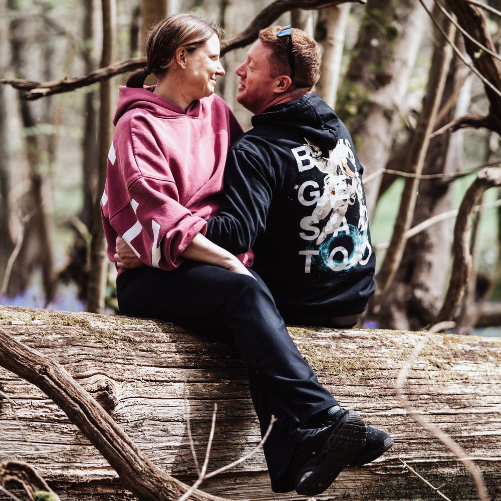 Couple sitting on a fallen log in a forest, wearing casual hoodies, sharing a romantic moment outdoors.