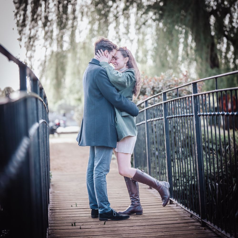 Couple sharing a romantic embrace on a wooden bridge surrounded by lush green weeping willow trees.