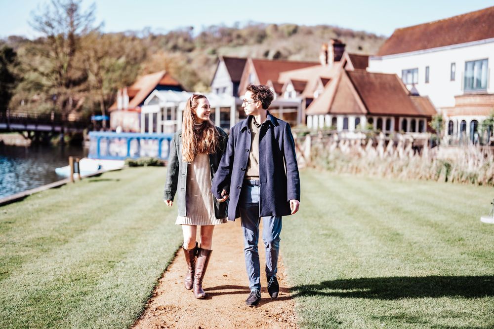 A couple walks hand-in-hand along a garden path beside a charming riverside estate on a sunny day.