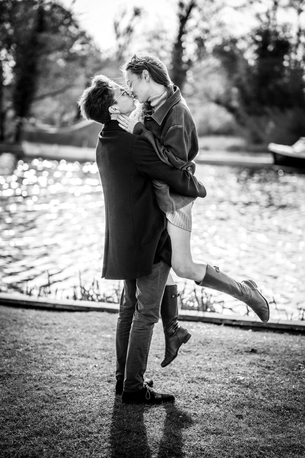 Black and white photo of a couple kissing by a pond, man lifting woman in a romantic outdoor embrace.