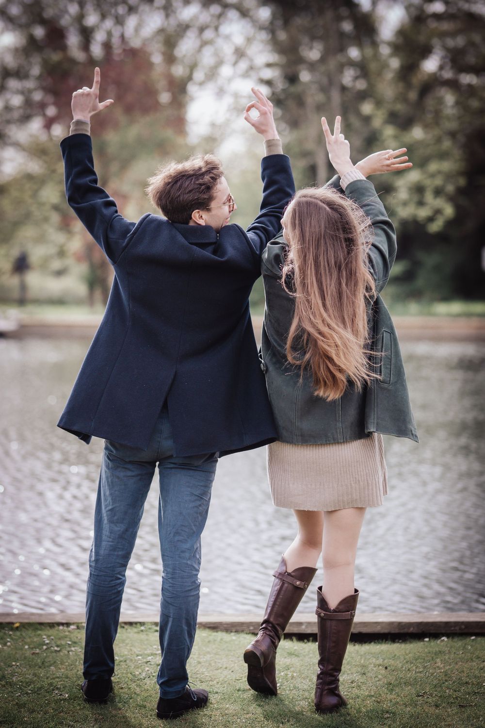 Couple standing by a lake with arms raised making hand gestures, viewed from behind in a park setting.