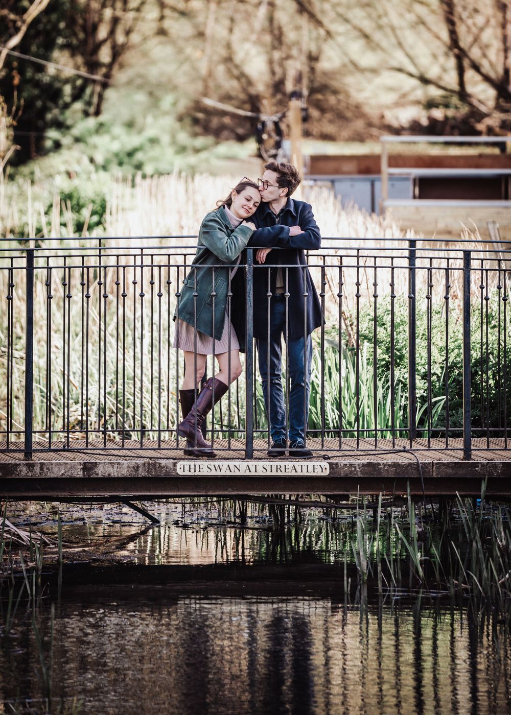 Couple sharing a romantic moment on a bridge over a pond at The Swan at Streatley, surrounded by lush greenery.