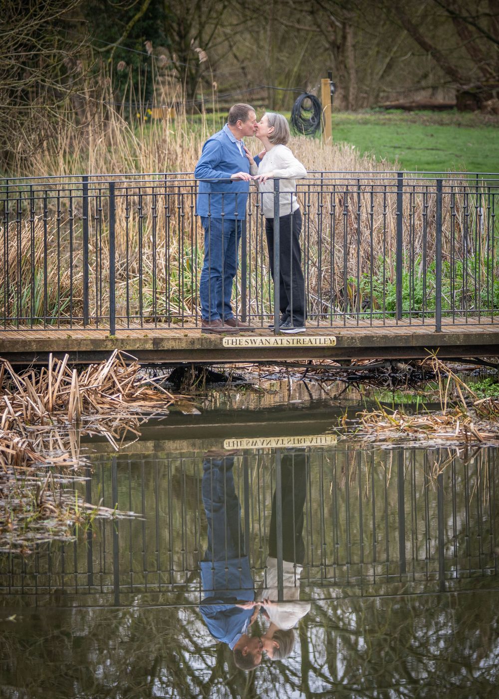 Couple sharing a romantic kiss on a wooden bridge over a calm reflective pond in a lush green park.