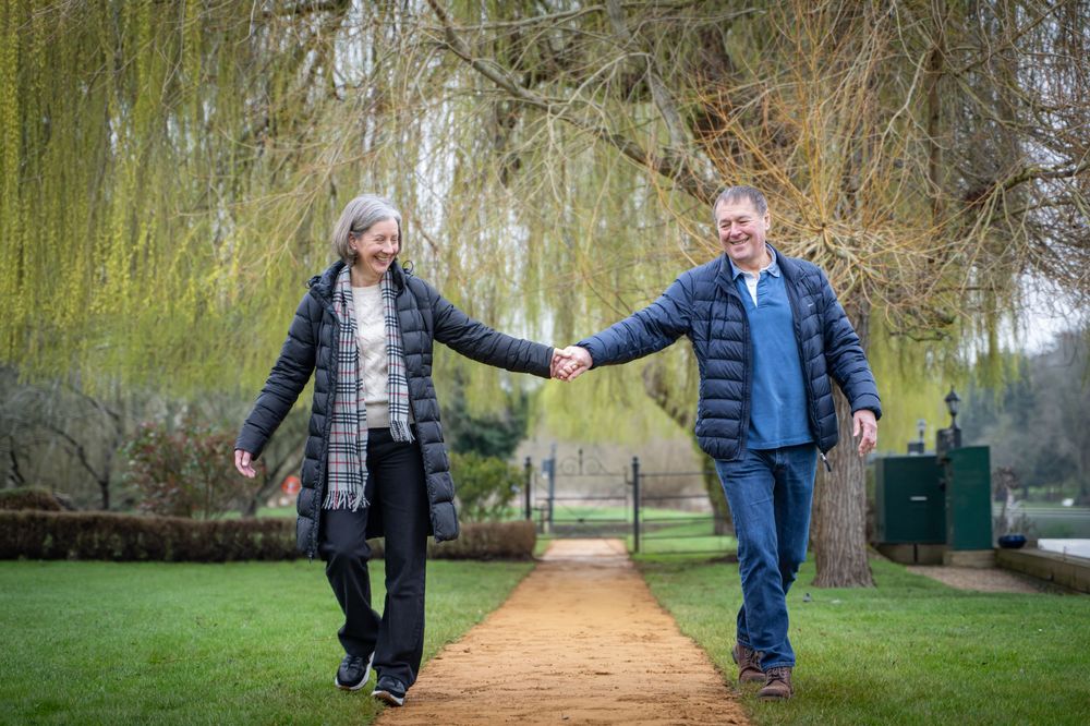 Elderly couple holding hands walking on a garden path under a weeping willow tree in a lush green park.