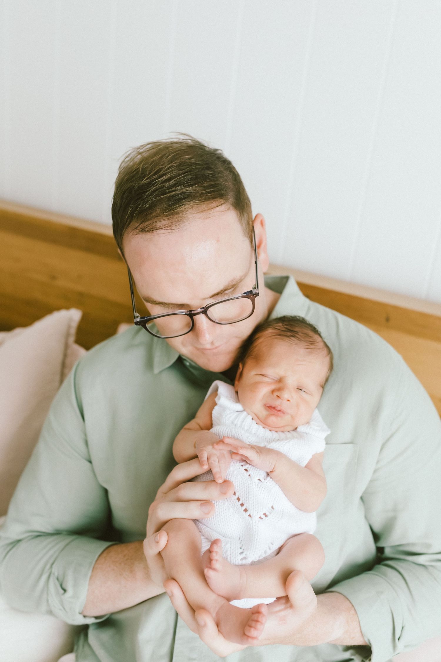 A father in a green shirt and glasses tenderly holds his newborn baby dressed in white lace.