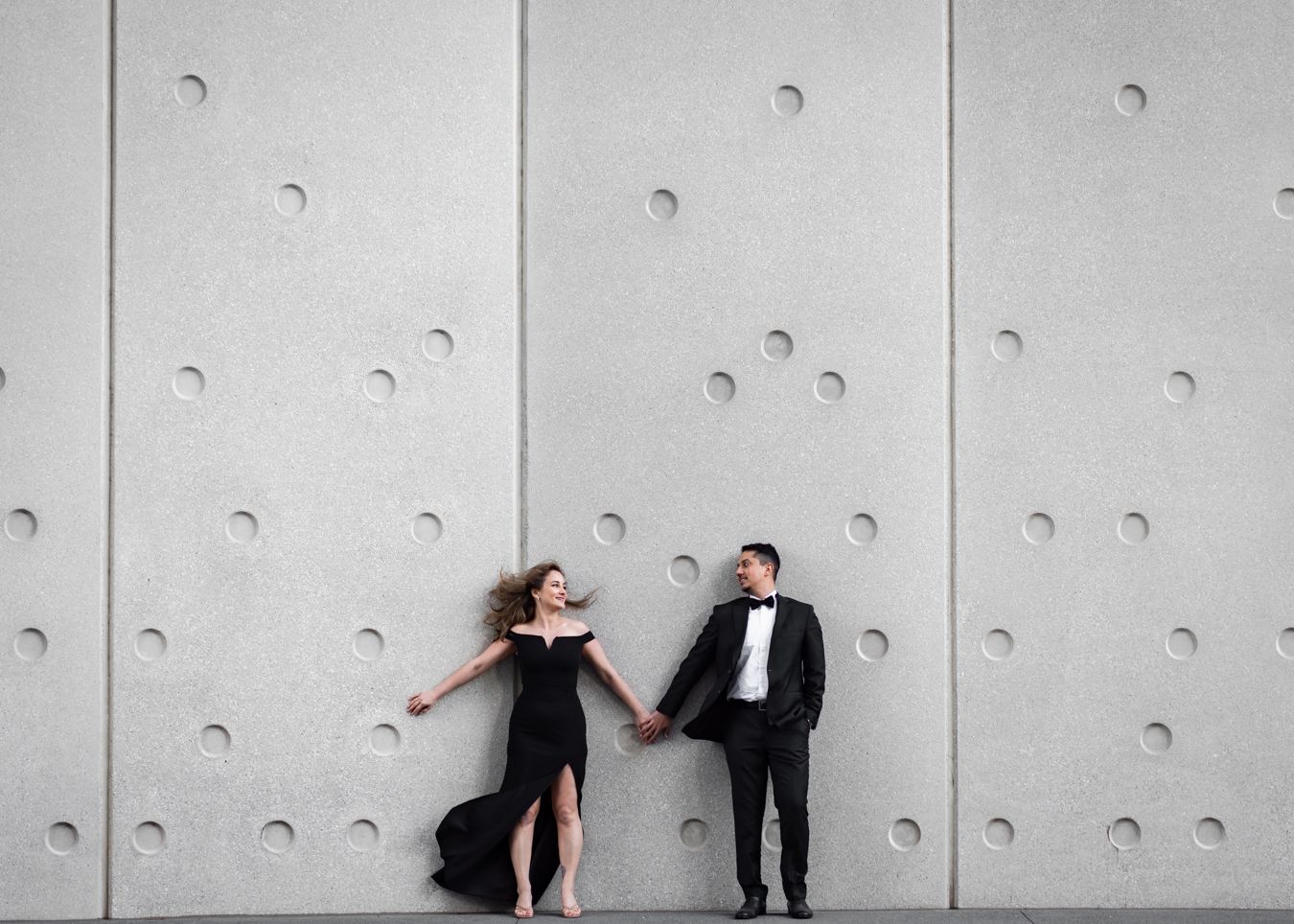A couple in formal evening wear dances together in black and white against a textured wall.