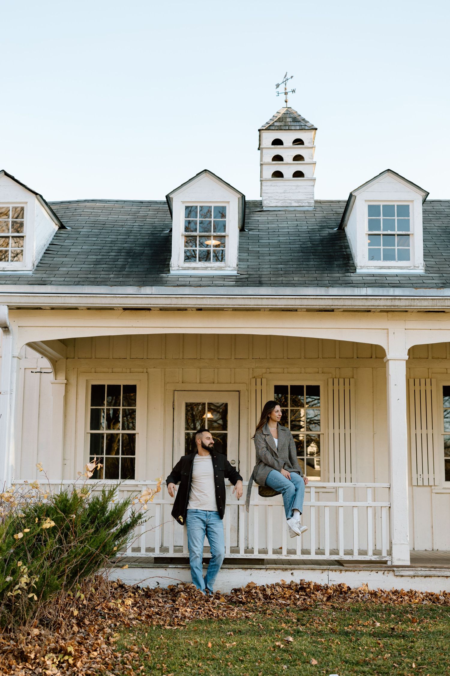 Couple posing in front of a charming white colonial farmhouse with dormers and a cupola on a fall day.