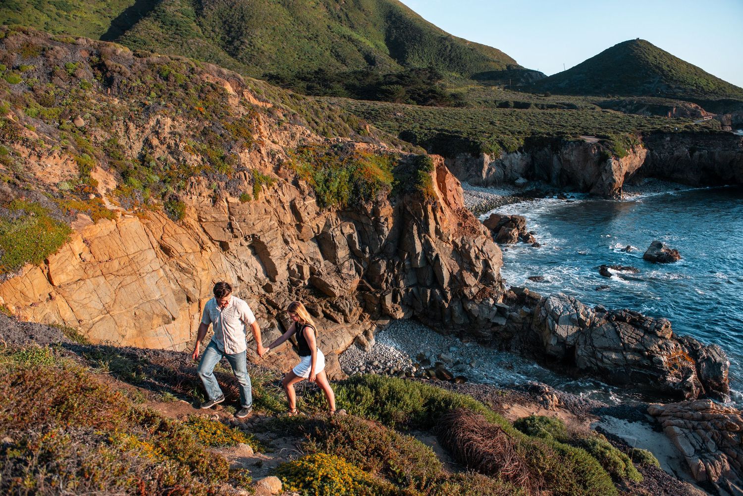 Two hikers exploring rocky coastal cliffs overlooking blue ocean waters with green hills in the background at sunset.