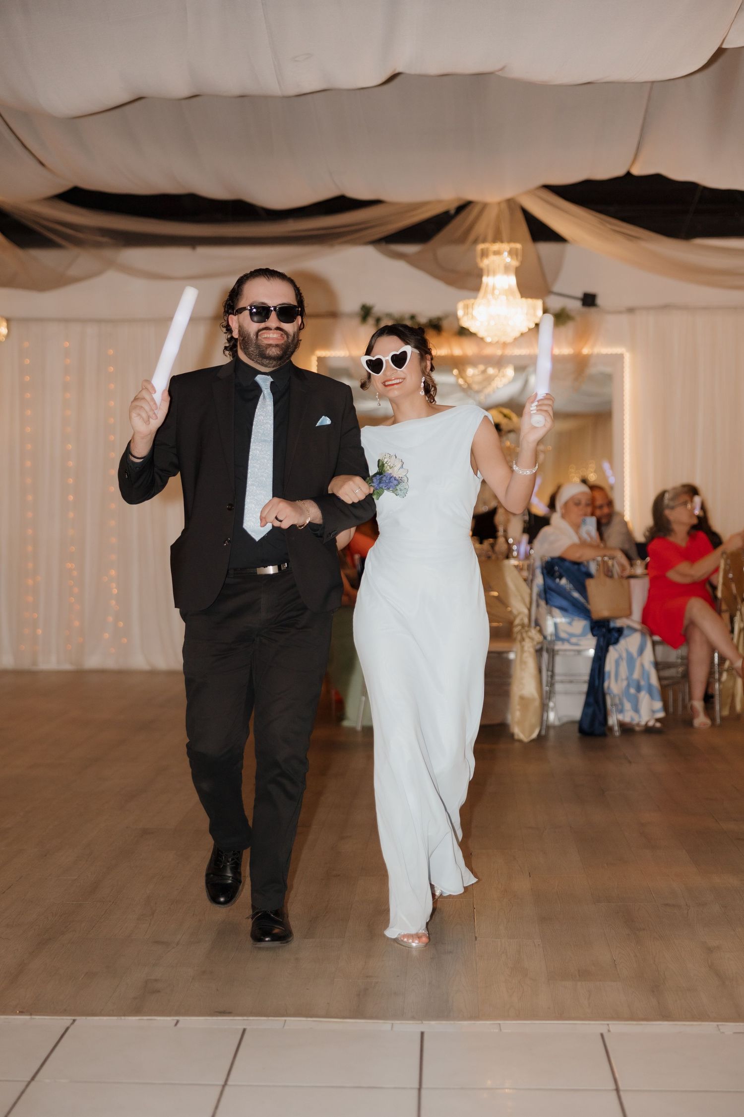 Bride and groom wearing sunglasses making grand entrance at wedding reception with glow sticks.
