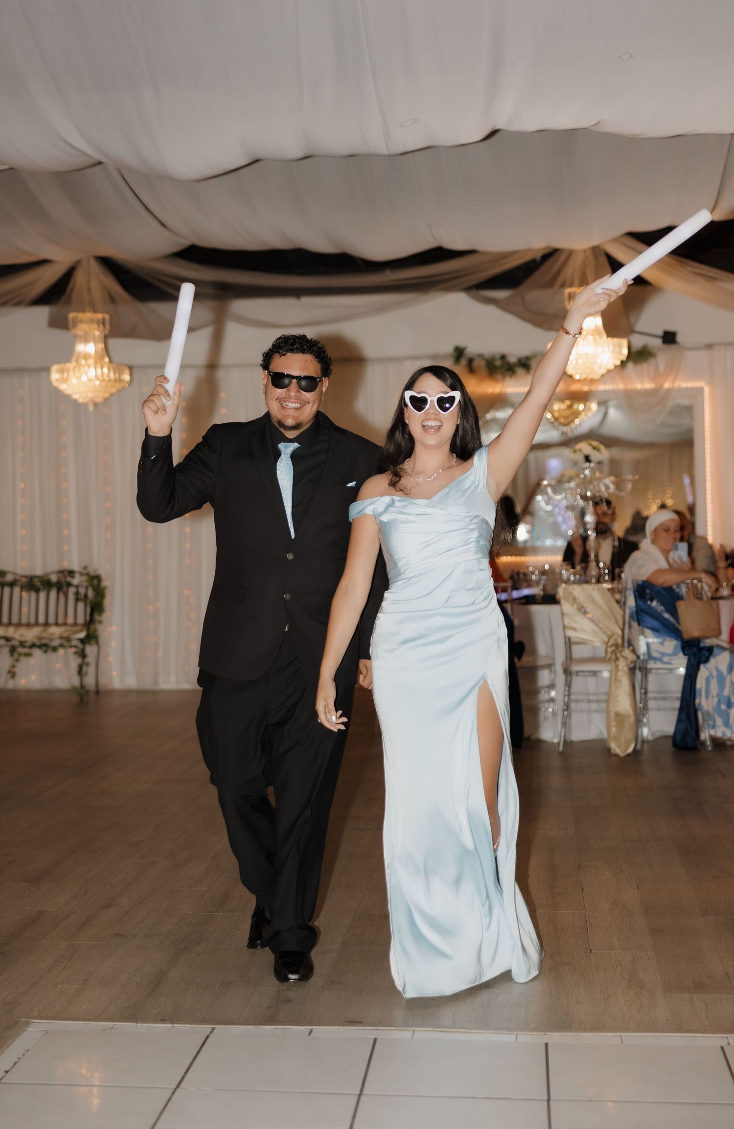 Couple in formal attire with sunglasses celebrating grand entrance at wedding reception.