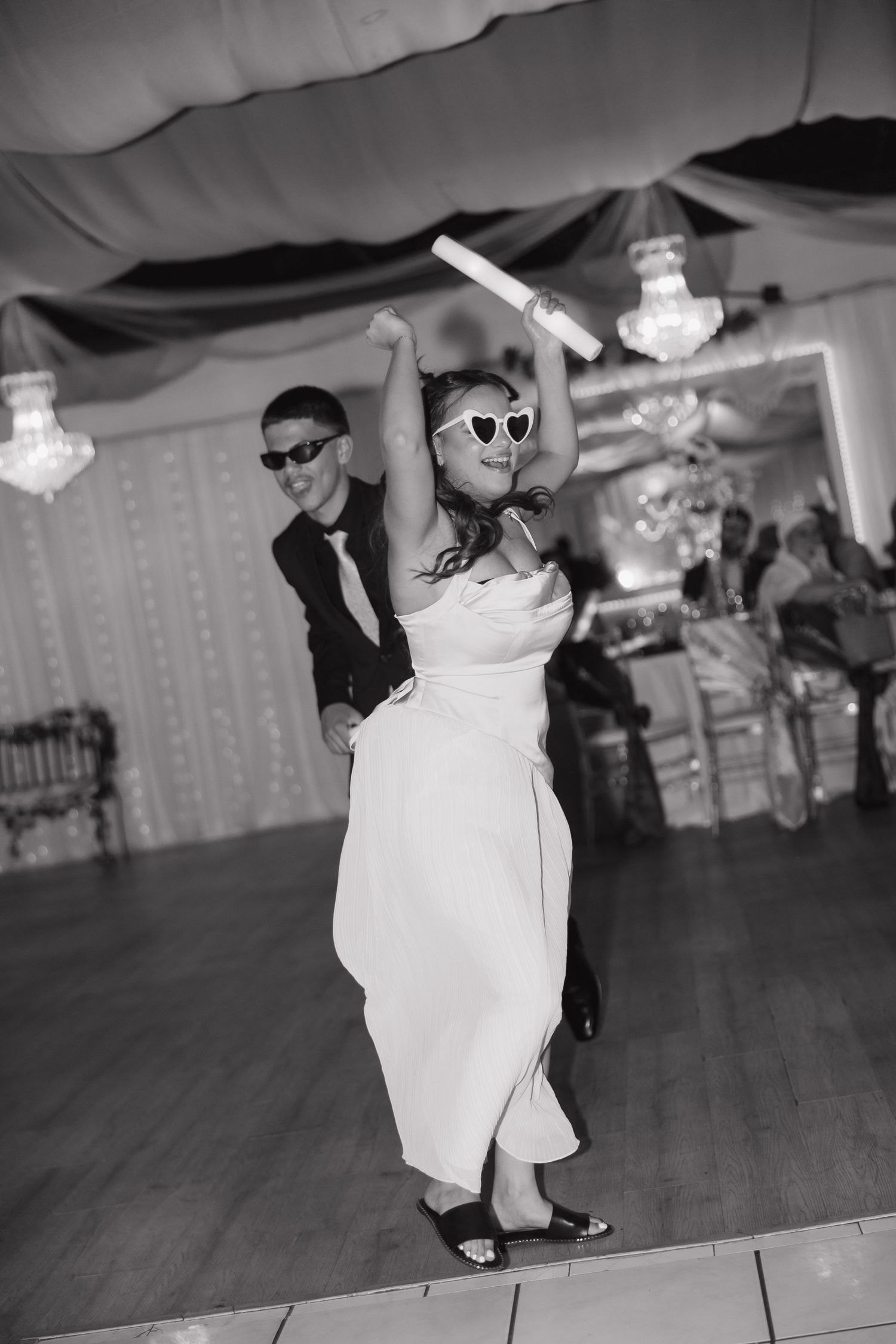 Black and white photo of bride and groom dancing at wedding reception wearing sunglasses under chandeliers.