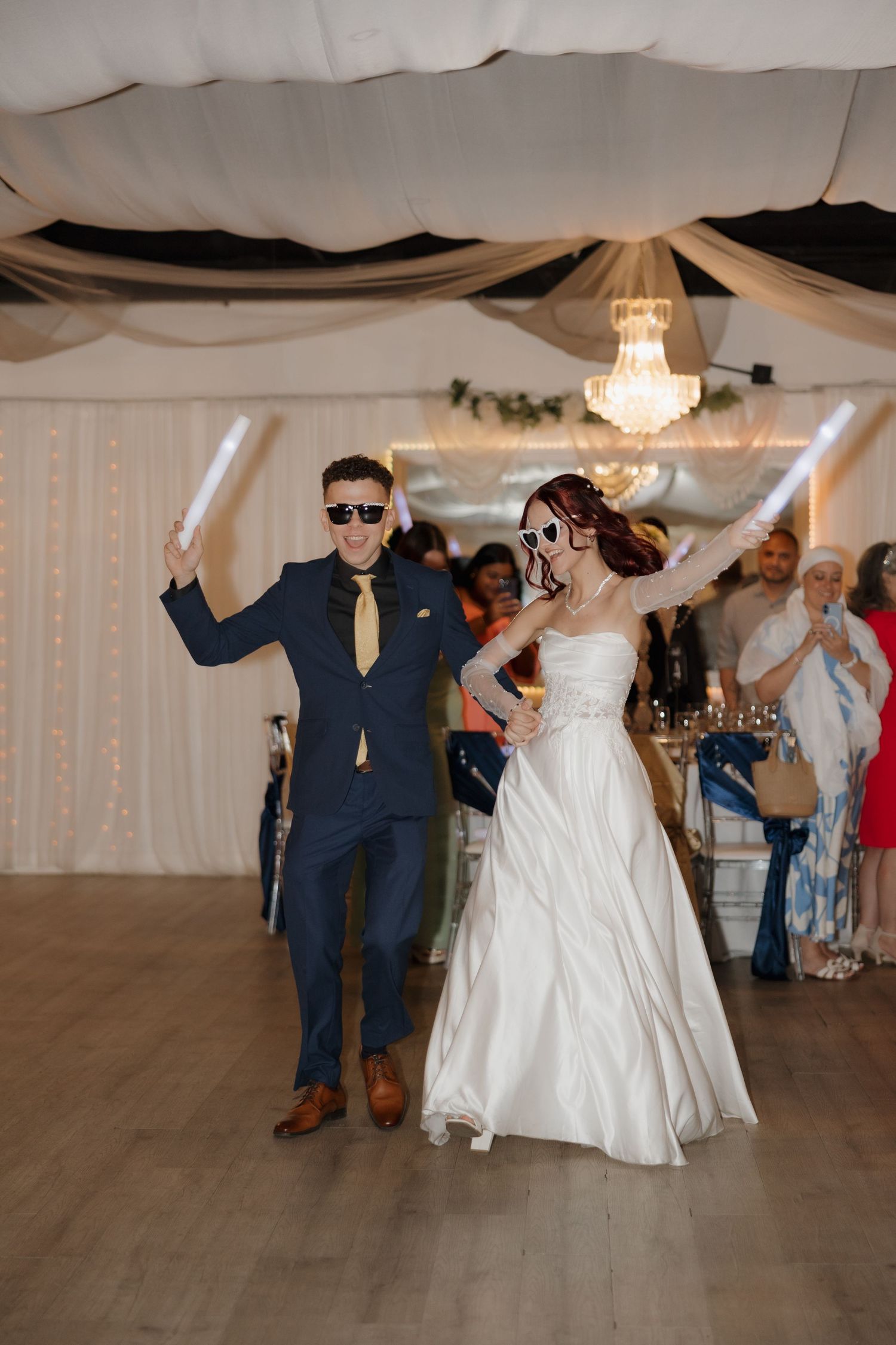 Bride and groom celebrate reception entrance wearing fun sunglasses while holding glowing foam sticks.