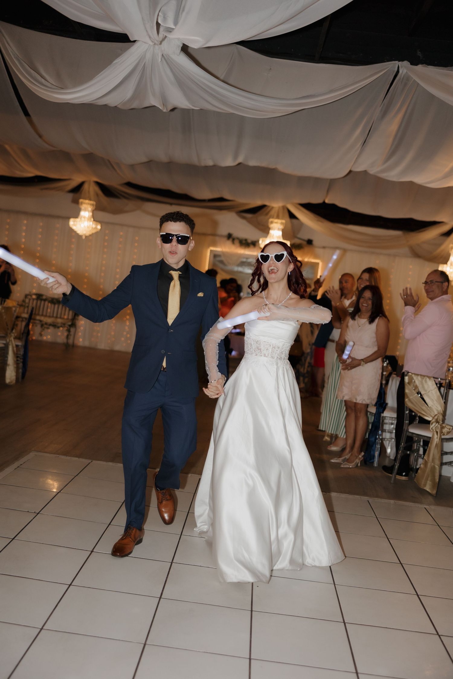 Bride and groom in sunglasses dancing at wedding reception under draped white ceiling decor.