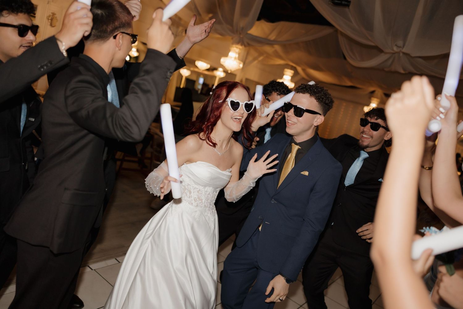 Bride in white strapless gown and groom in navy suit wear heart sunglasses celebrating at wedding reception with guests.