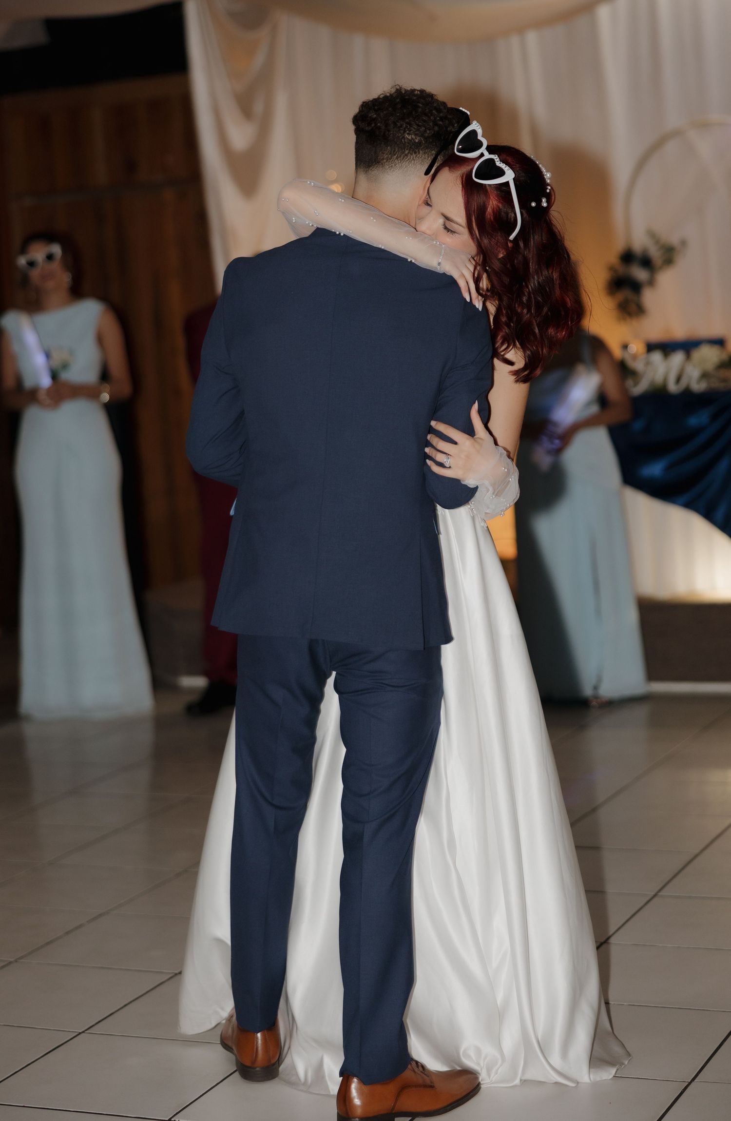 Bride and groom share first dance at wedding reception, bridesmaids in blue gowns visible in background.