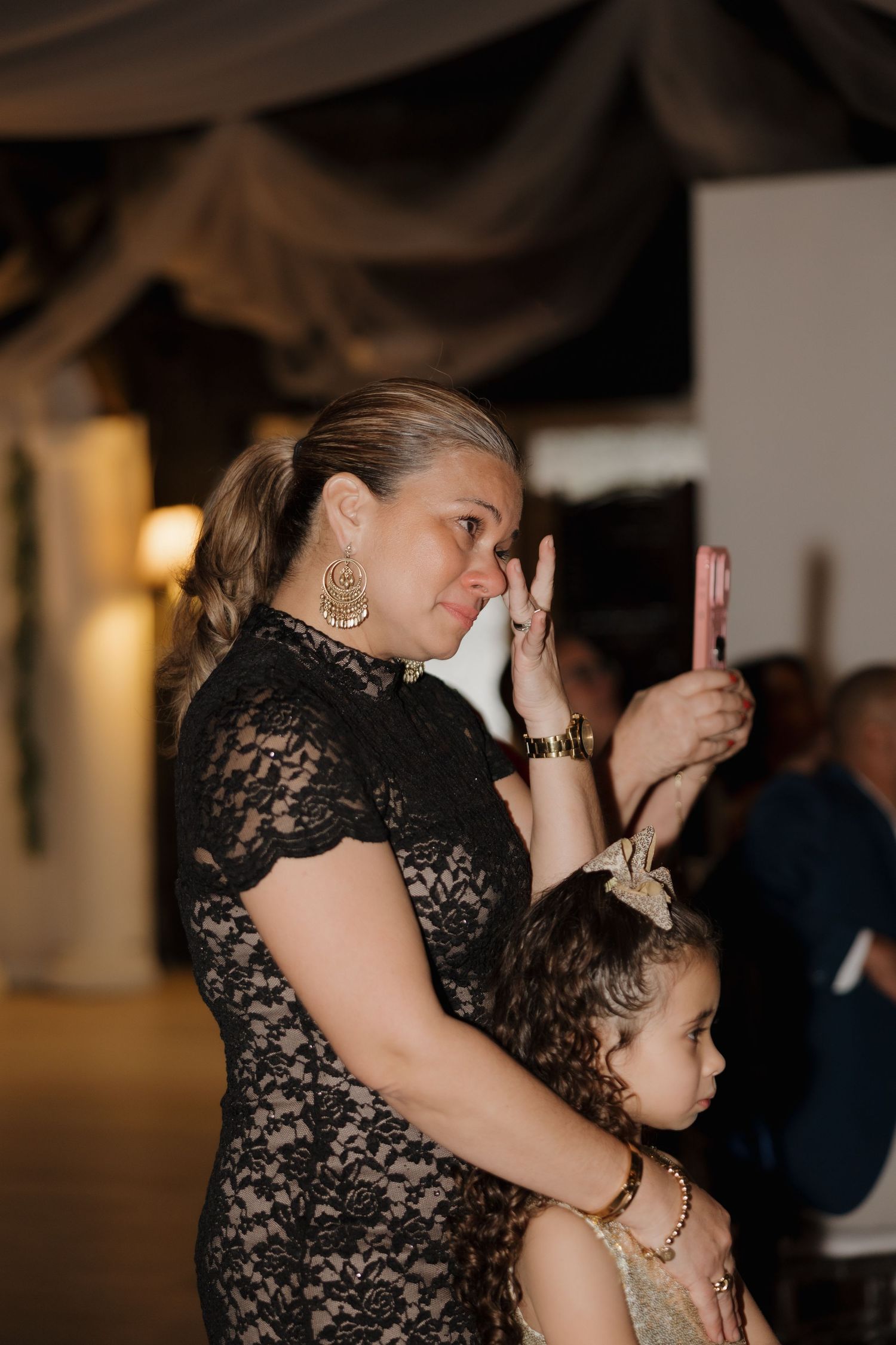 Emotional wedding guest in black lace dress wipes tears while holding child at reception.