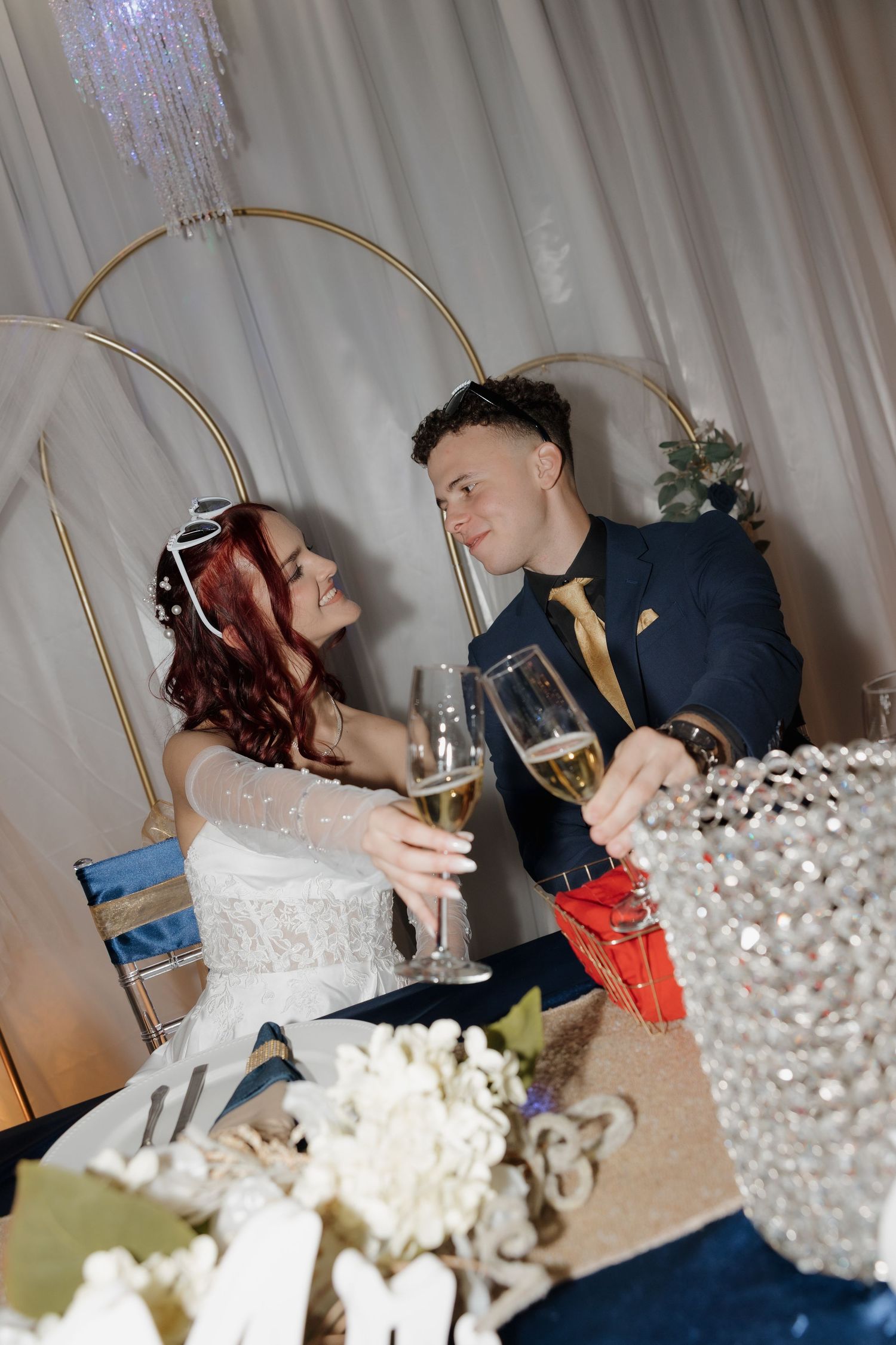 Bride and groom toasting with champagne glasses at sweetheart table during elegant wedding reception.